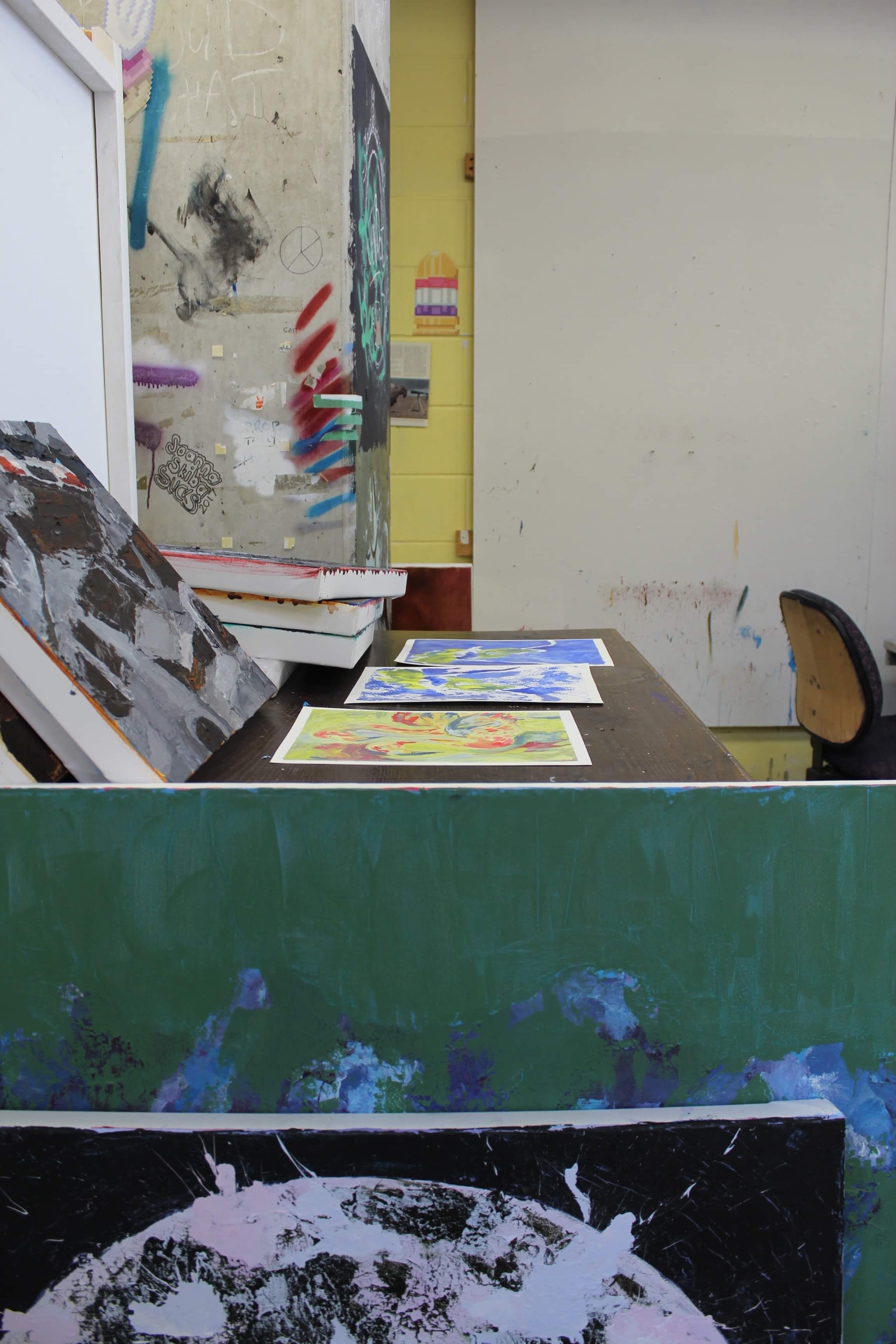 Photo of the artist's studio with paintings spread across a desk for review.